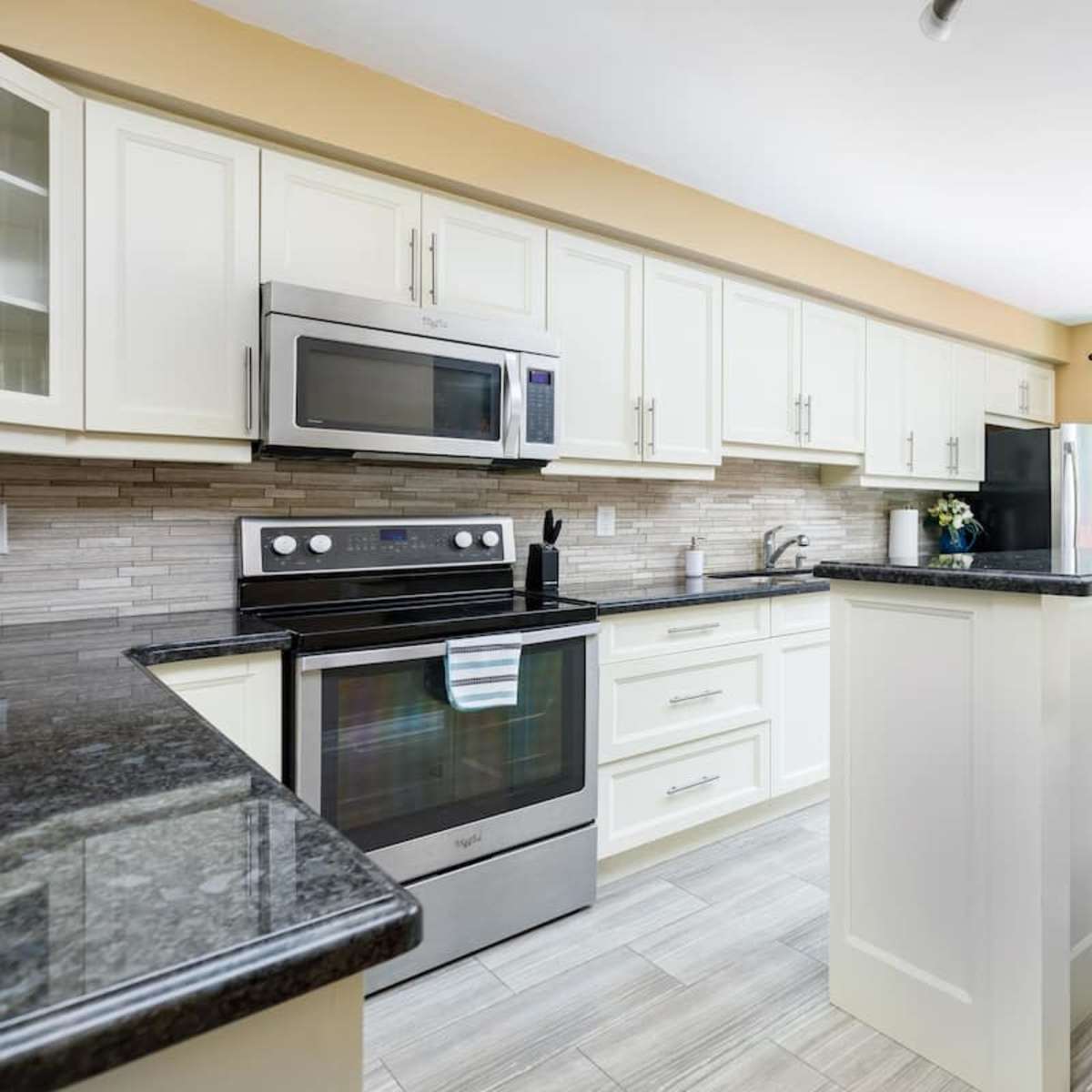 A modern kitchen features light cabinets, a dark countertop, stainless steel appliances, and a seating area with bar stools.