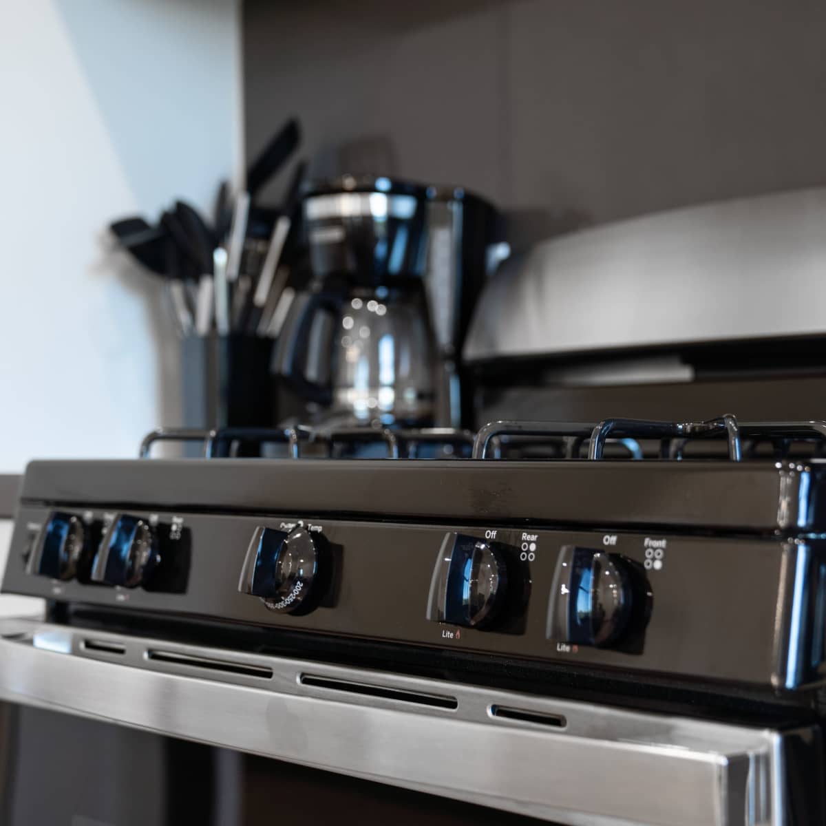 A black gas stove is positioned next to a silver countertop and kitchen utensils, with a coffee maker in the background.