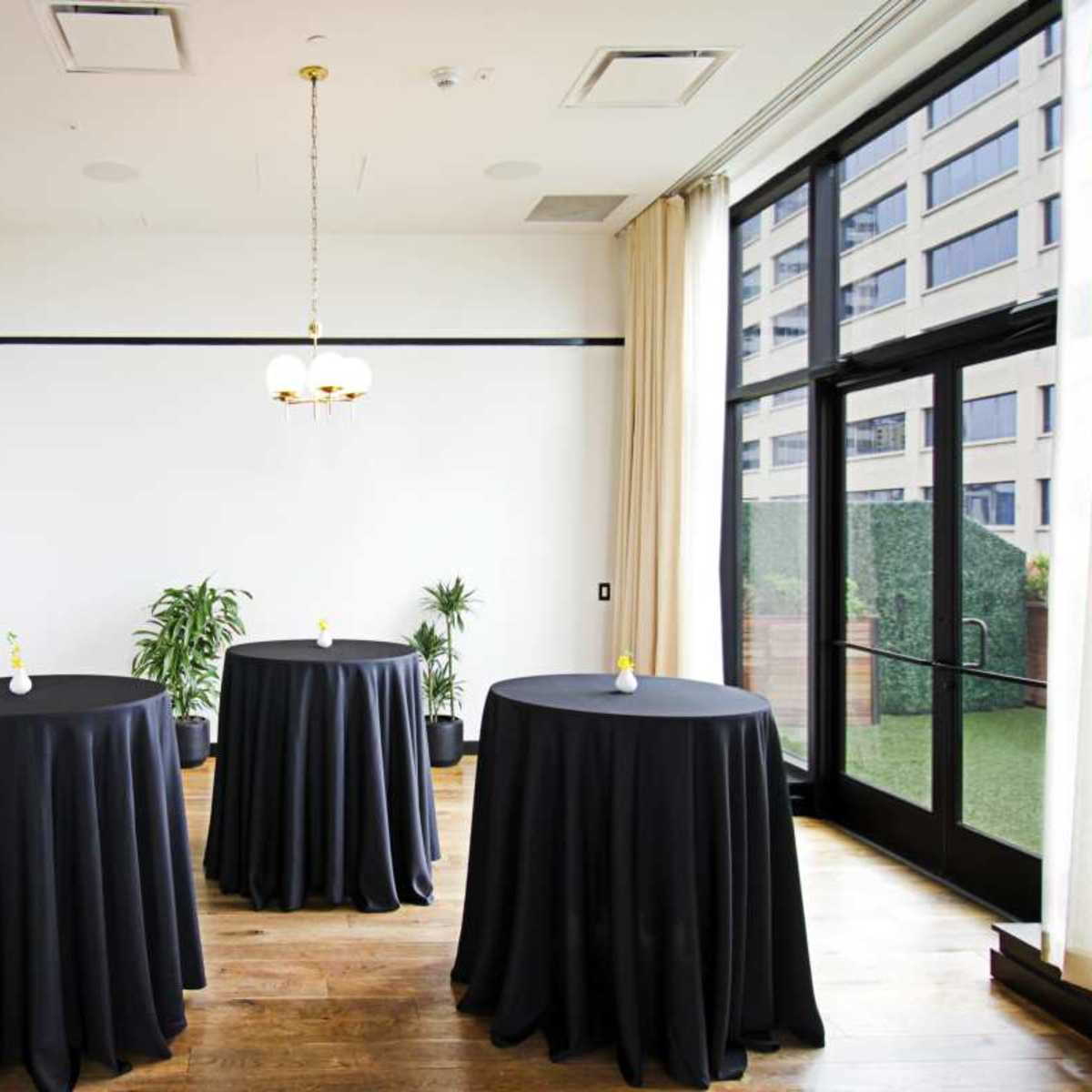 A room with three round tables covered in black tablecloths, situated near large glass doors that lead to an outdoor area.