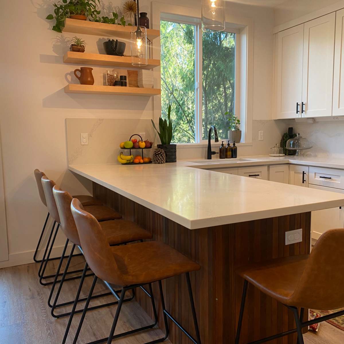 A modern kitchen features a large island with bar stools, a window overlooking greenery, and neatly arranged kitchenware and produce.