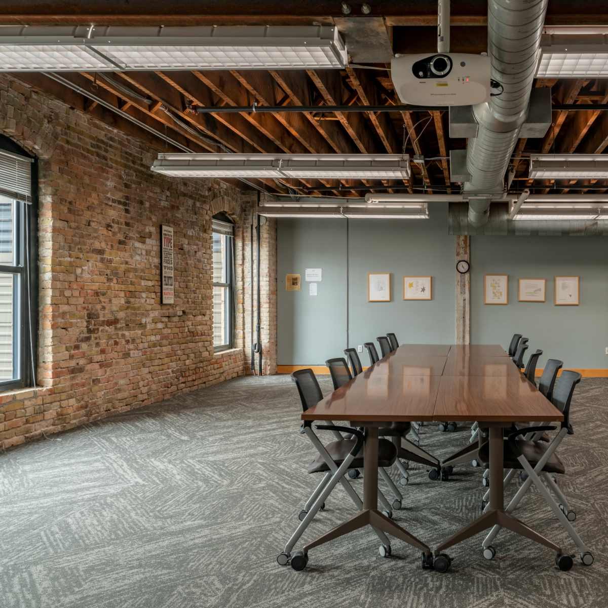 A spacious conference room featuring a long wooden table with rolling chairs, brick walls, and large windows.