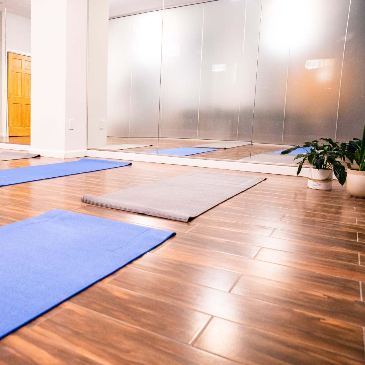 An empty yoga studio with blue mats arranged on a wooden floor and mirrors reflecting the space.