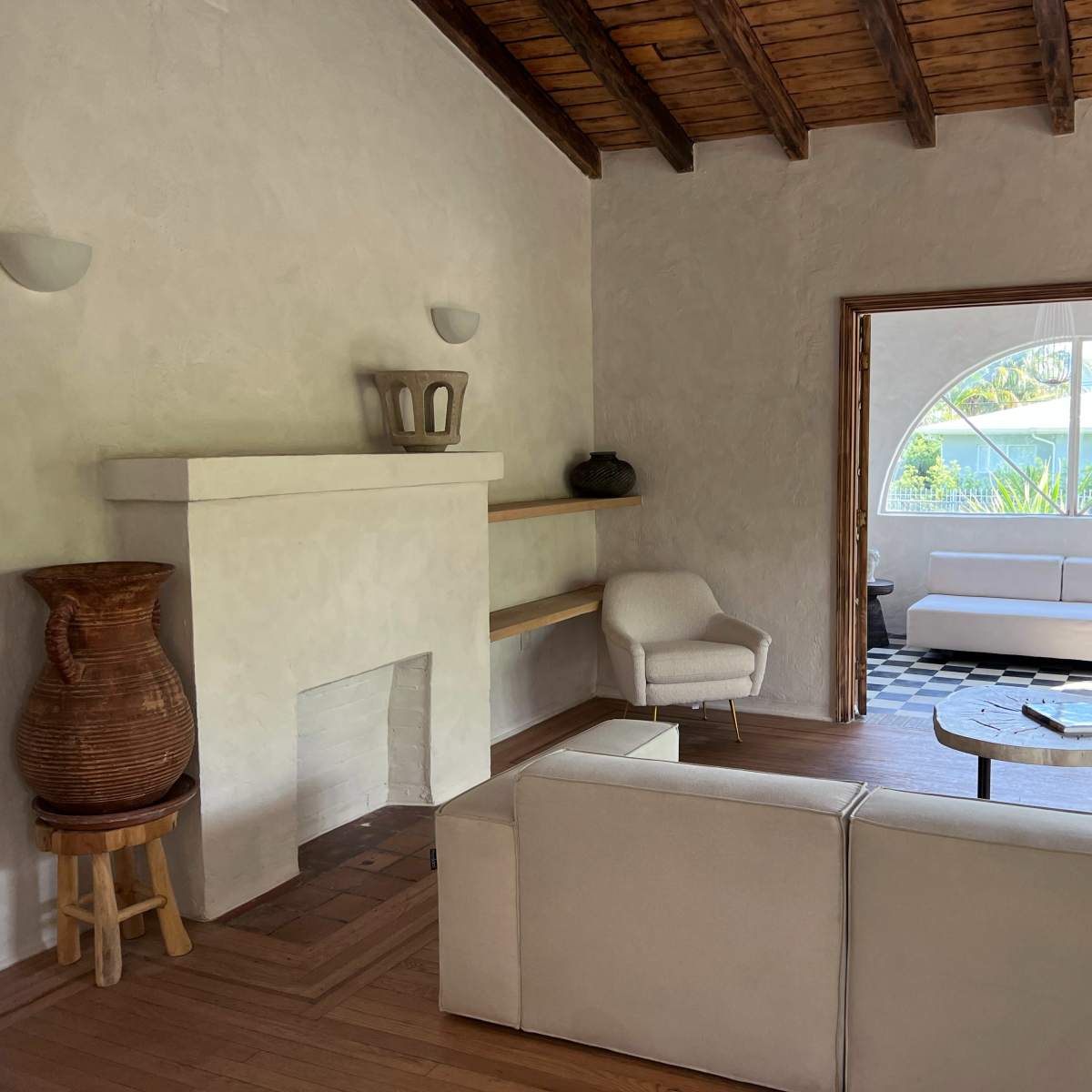 A living room with a white sofa, a wooden ceiling, and a fireplace, leading to an outdoor area visible through an arched window.