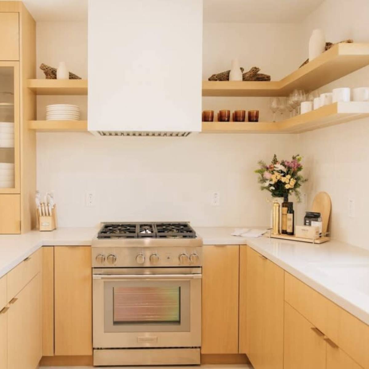A modern kitchen features light wood cabinets, a stainless steel stove, and open shelves displaying dishes and decorative items.