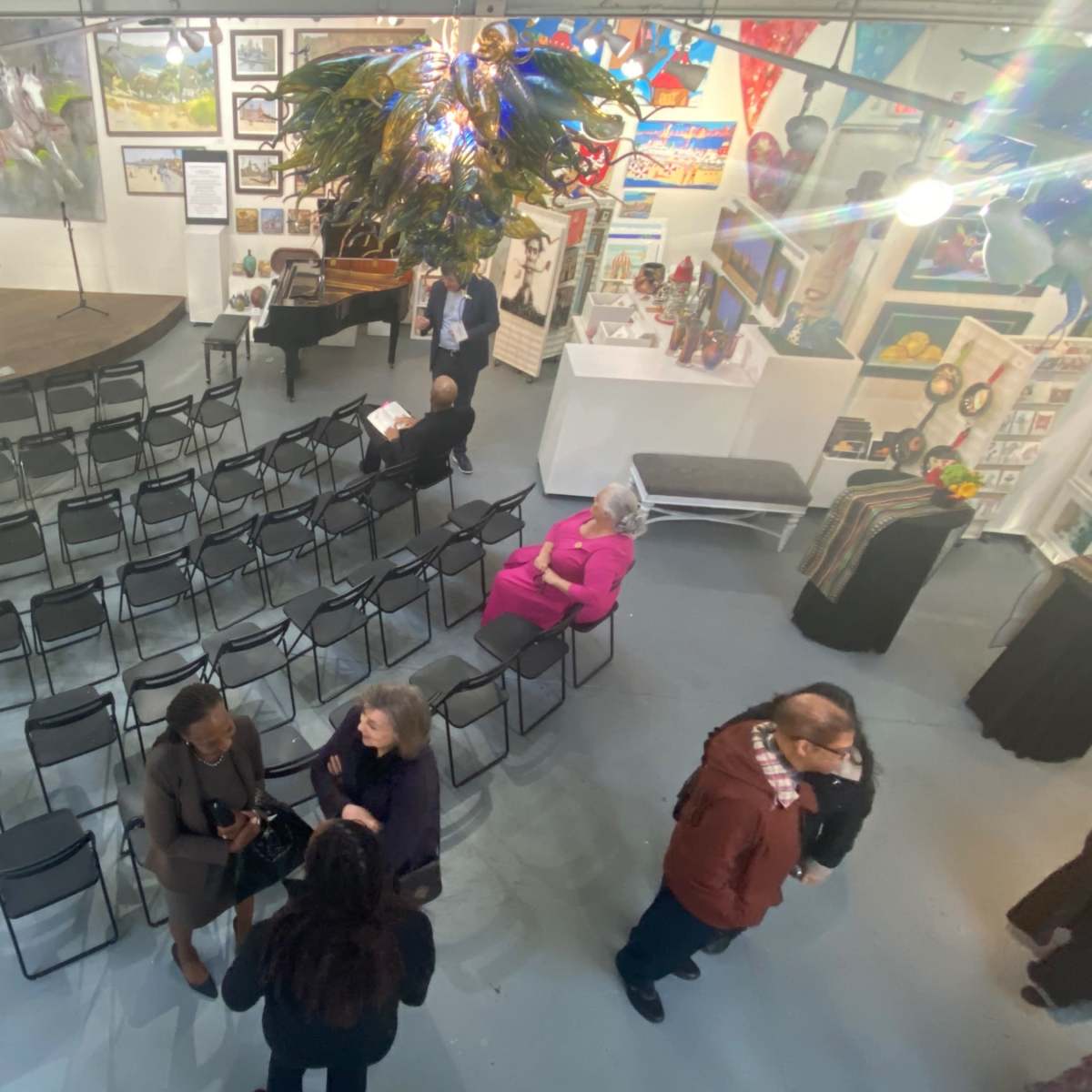 An event space with rows of black chairs set up for an audience, a stage at the front, and several people mingling in various areas of the room.