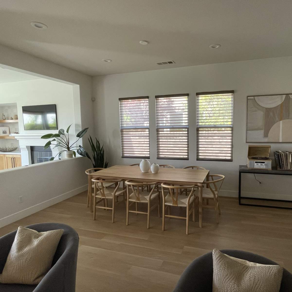 A modern dining area features a wooden table with chairs, positioned near large windows with blinds, beside a console table and artwork on the wall.