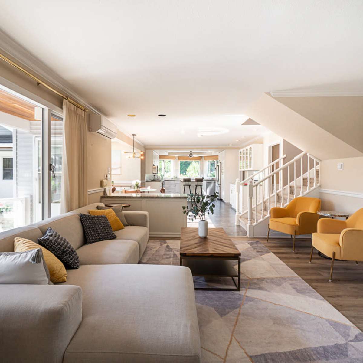 The living room features a light-colored sectional sofa, a wooden coffee table, and two orange armchairs, leading to an open kitchen visible in the background.