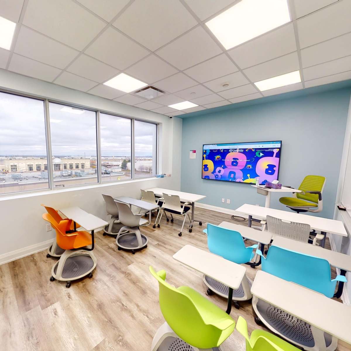 A modern classroom with colorful chairs arranged around small tables, equipped with a large screen displaying animated graphics and large windows offering an outside view.