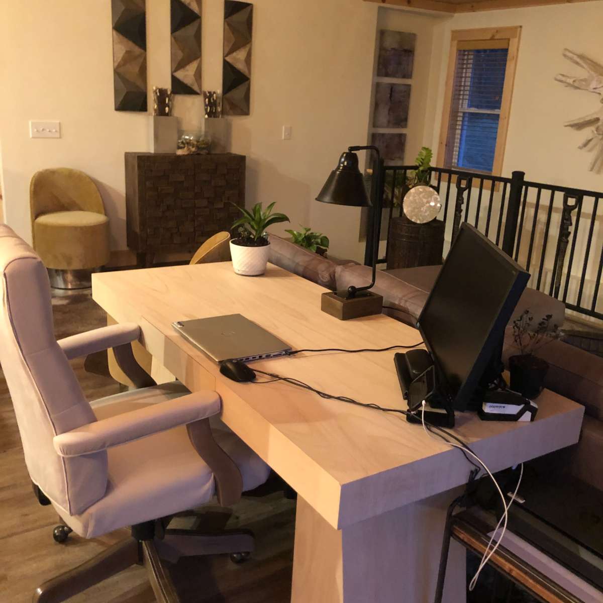 A modern workspace featuring a light wood desk, an office chair, a computer monitor, and decorative plants, set in a room with wooden accents and artwork on the walls.