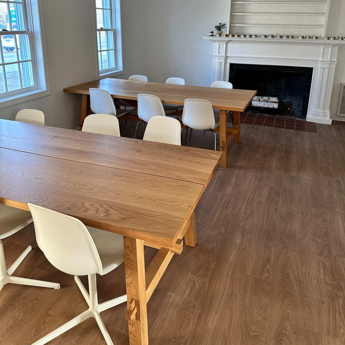A bright room with two wooden tables surrounded by white chairs, and a fireplace with shelves above it.