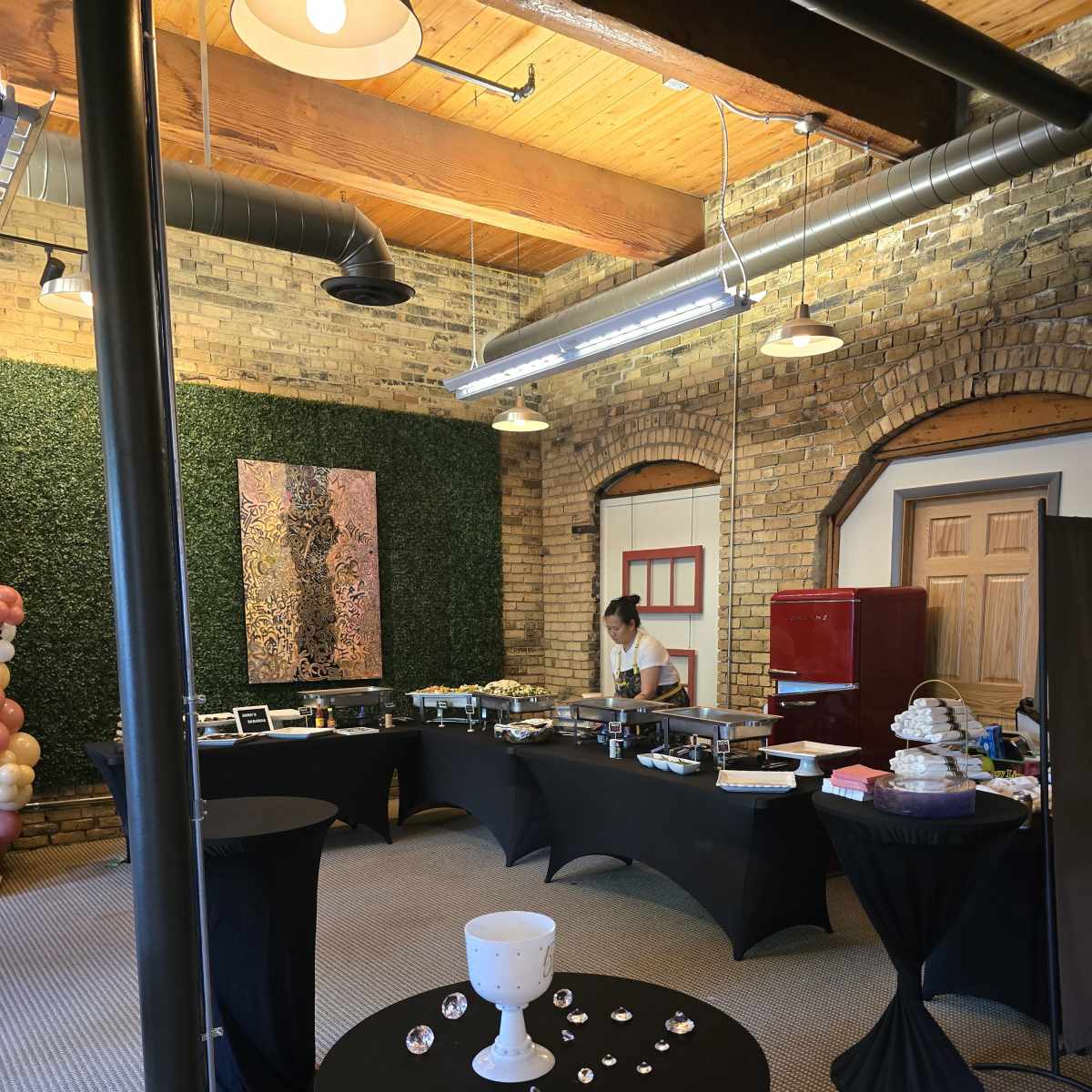 A spacious event room with black tablecloths, a buffet setup, and a person preparing food against a backdrop of brick walls and decorative greenery.