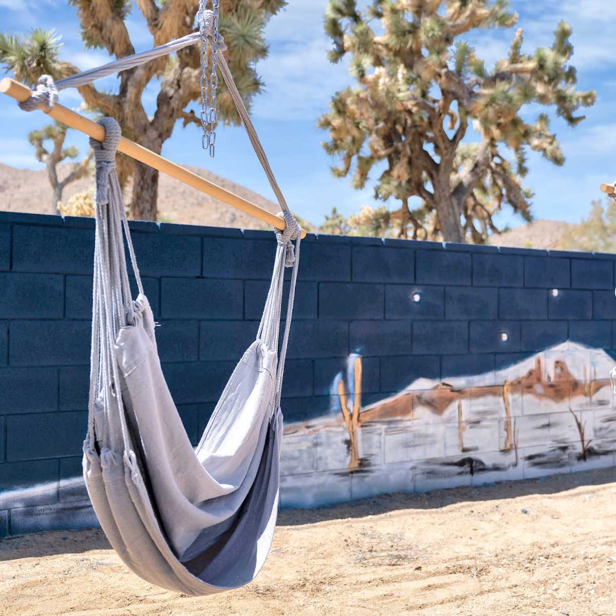 Two gray hammock swings hang from a wooden frame in a desert landscape with a mural of mountains on a black wall in the background.