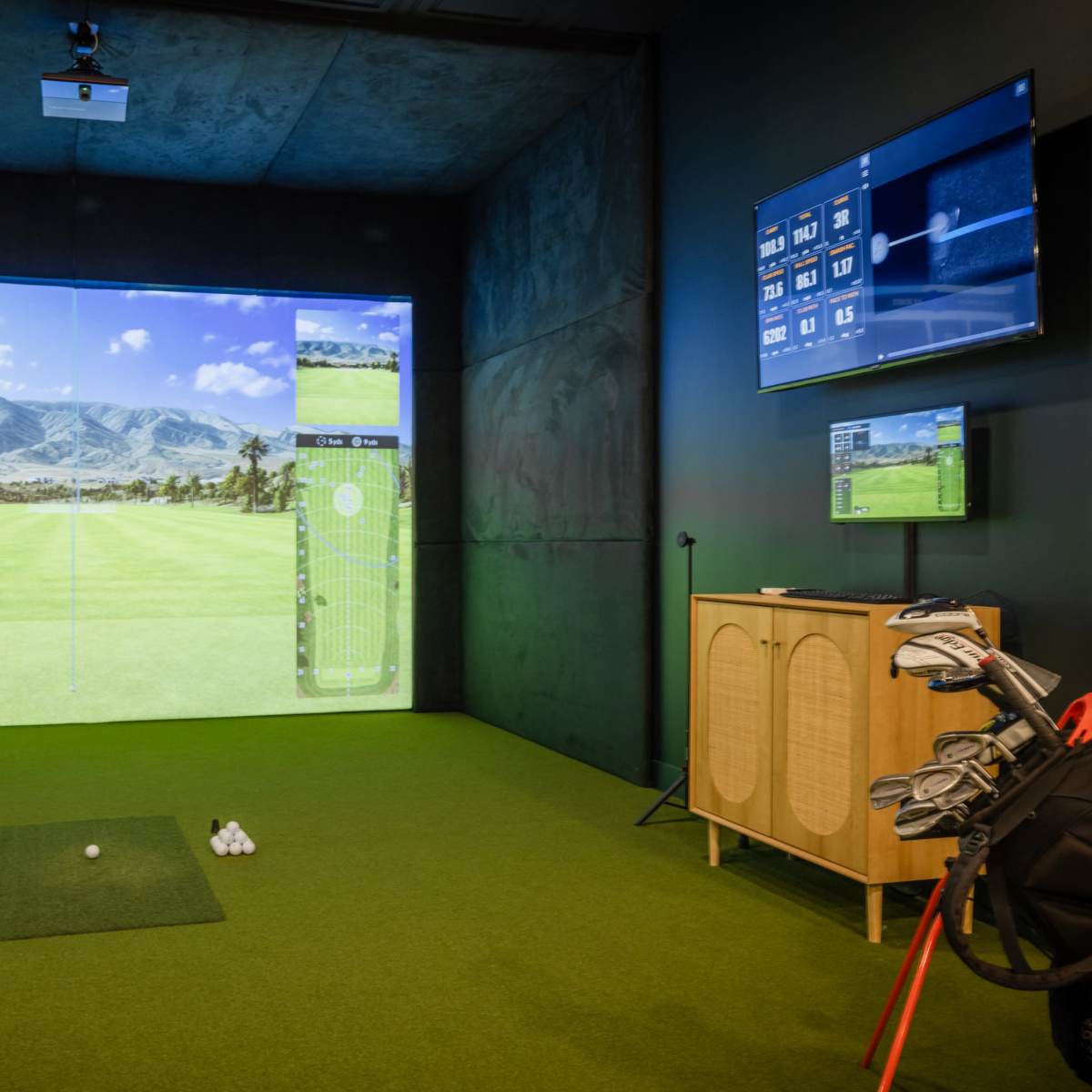 An indoor golf simulator setup with a large projection screen displaying a golf course, a golf tee with balls, and a set of clubs beside a cabinet.