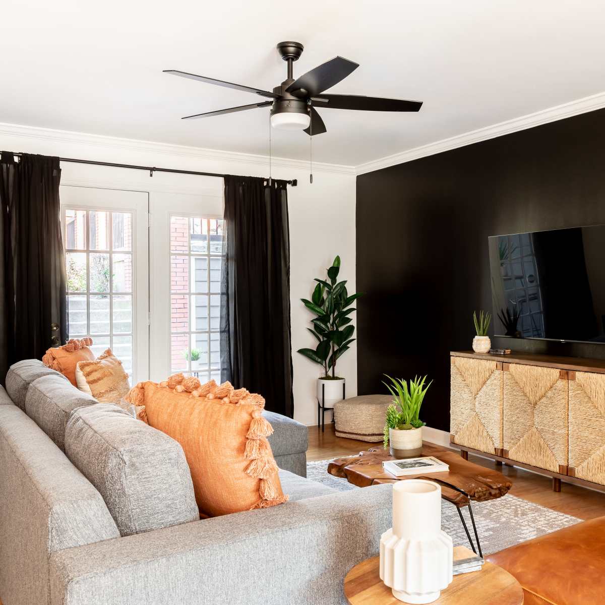 A modern living room features a gray sofa with orange cushions, a wooden media console, a TV mounted on a black wall, and potted plants near a door with black curtains.