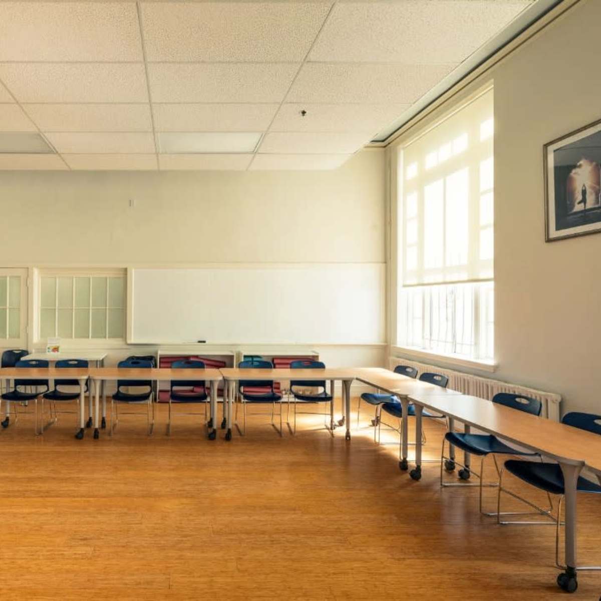 A sparsely furnished classroom with a long table and several chairs arranged around it, along with a whiteboard and a large window providing natural light.