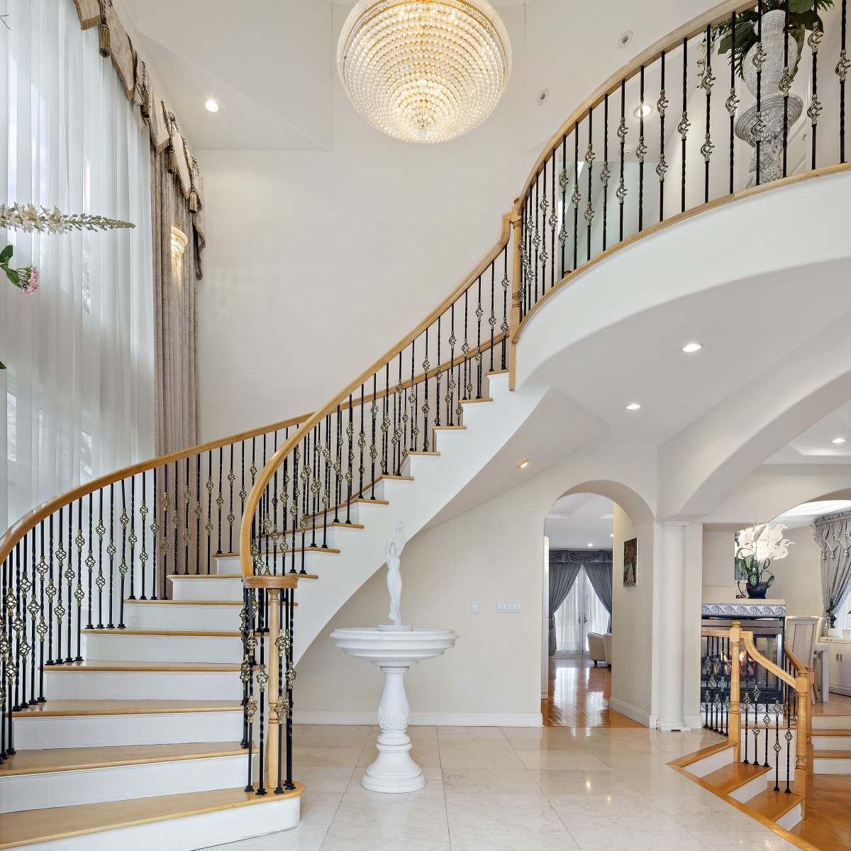 A spacious entryway with a curved wooden staircase, ornate railings, and a large chandelier overhead.