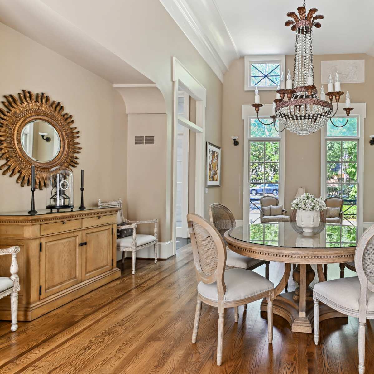 A dining room features a round wooden table surrounded by upholstered chairs, with a large chandelier overhead and windows allowing natural light to fill the space.