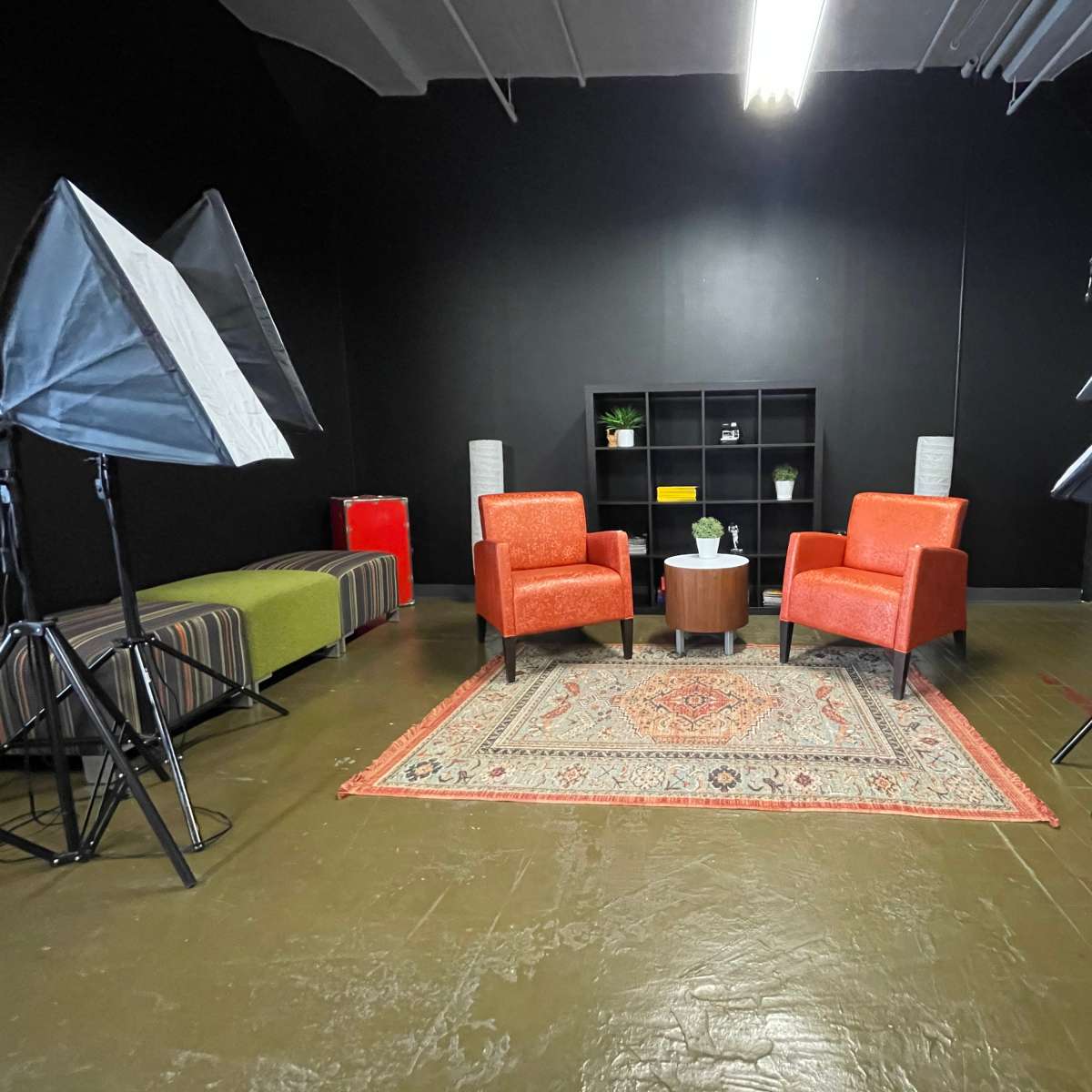 A studio setup featuring two orange chairs, a small round table, and multiple softbox lights arranged around a patterned rug on a dark wall background.