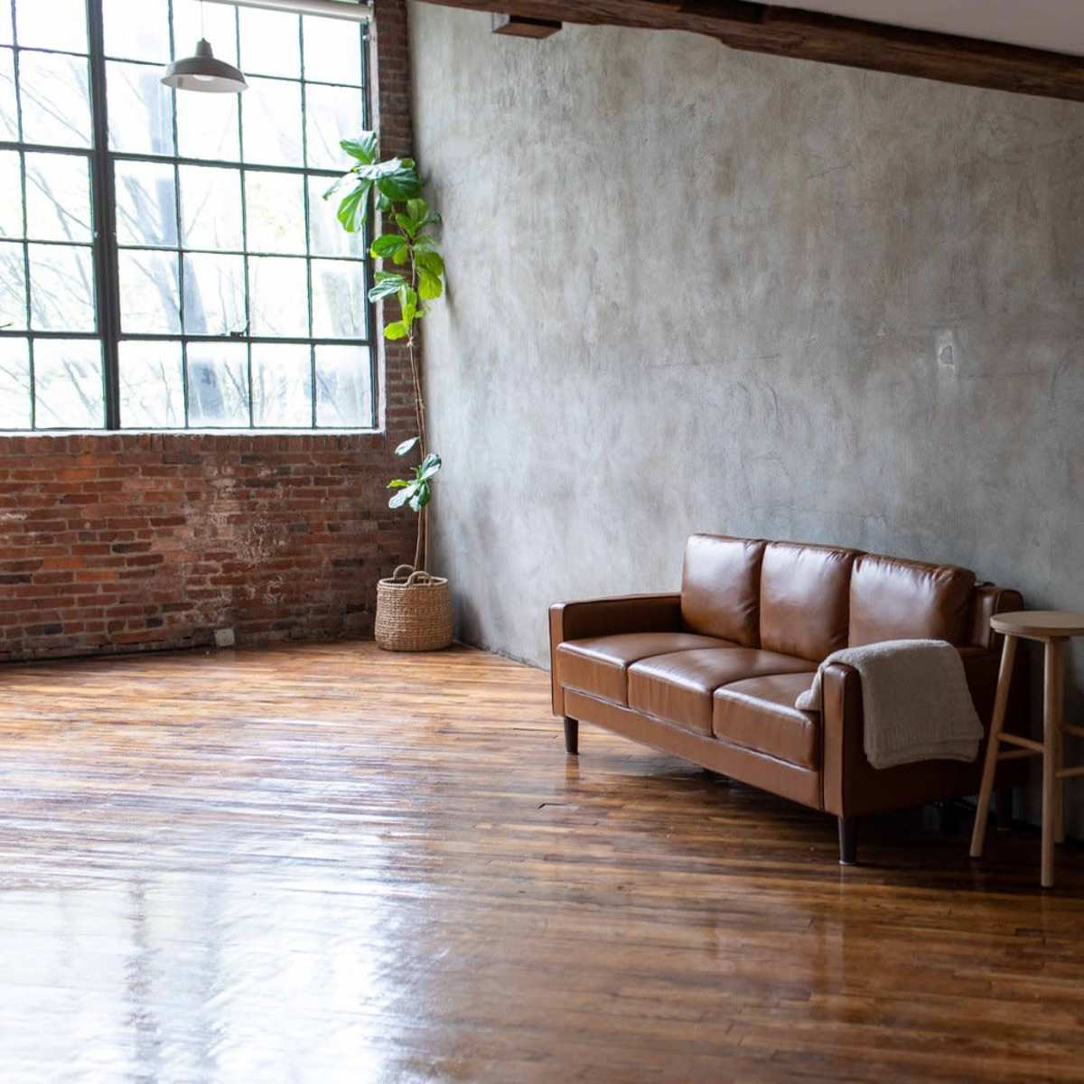 A spacious room with a brown leather sofa, a small wooden stool, a large mirror, and a concrete wall, complemented by a potted plant near a large window.