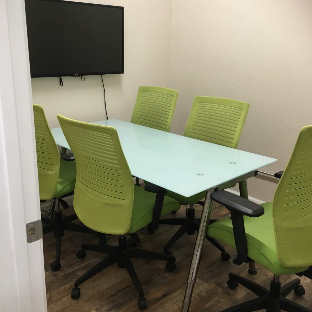 A glass conference table surrounded by six green chairs sits in a small, well-lit meeting room with a wall-mounted screen.