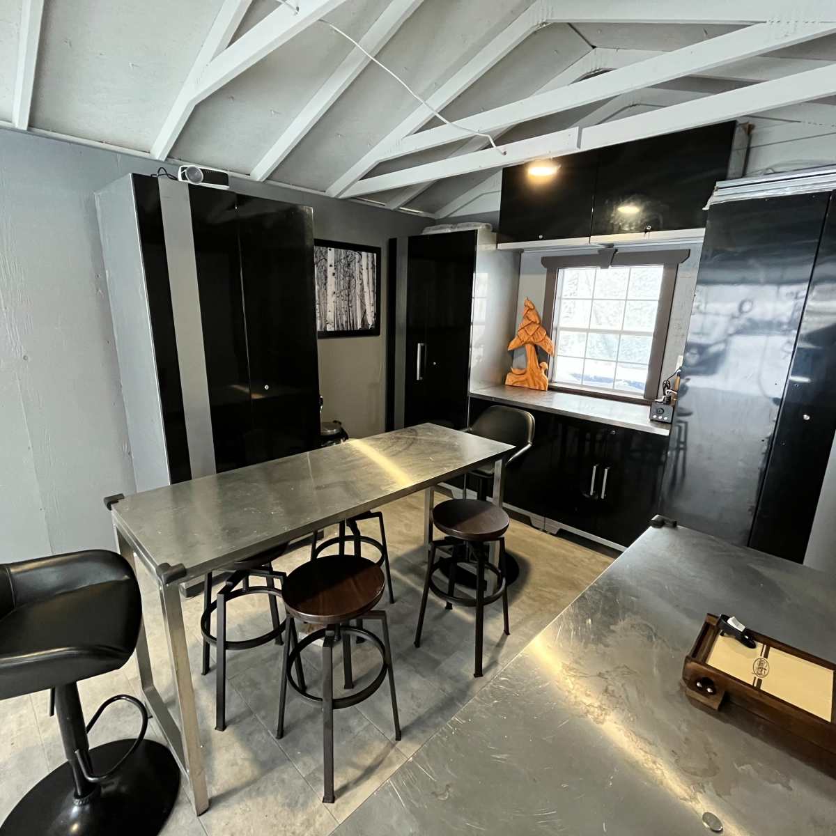 A modern kitchen features sleek black cabinets, a metallic table, and bar stools under a slanted ceiling with exposed beams.