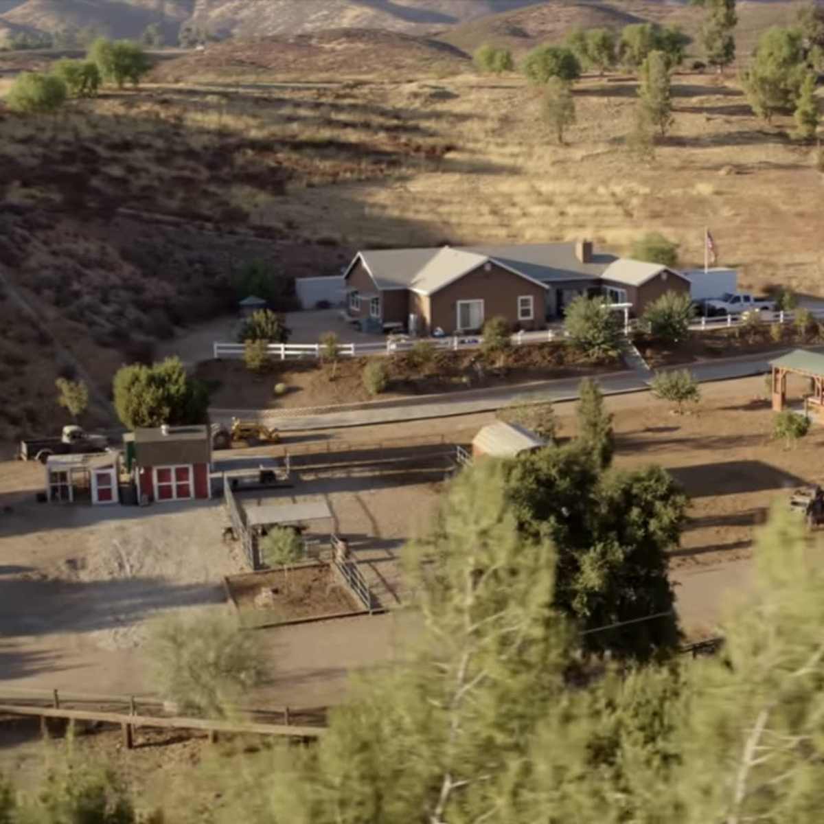 A rural property consisting of a house, several outbuildings, and fenced areas, surrounded by hills and greenery.