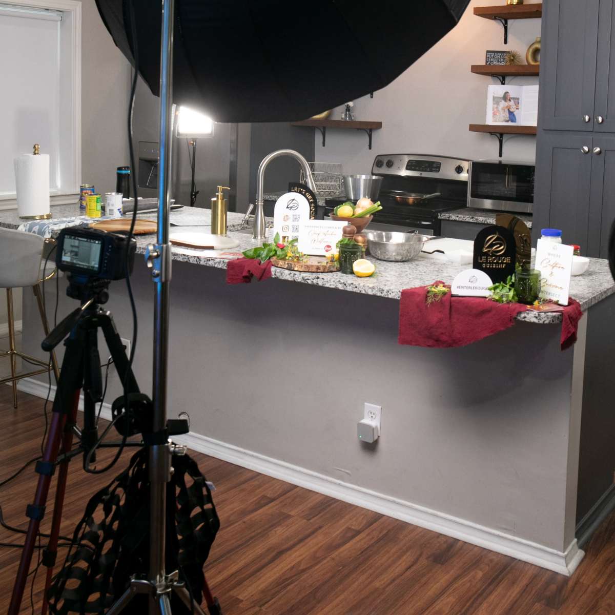 A kitchen setup for a photography session, featuring a black umbrella light over a granite countertop with various cooking ingredients and a camera on a tripod.