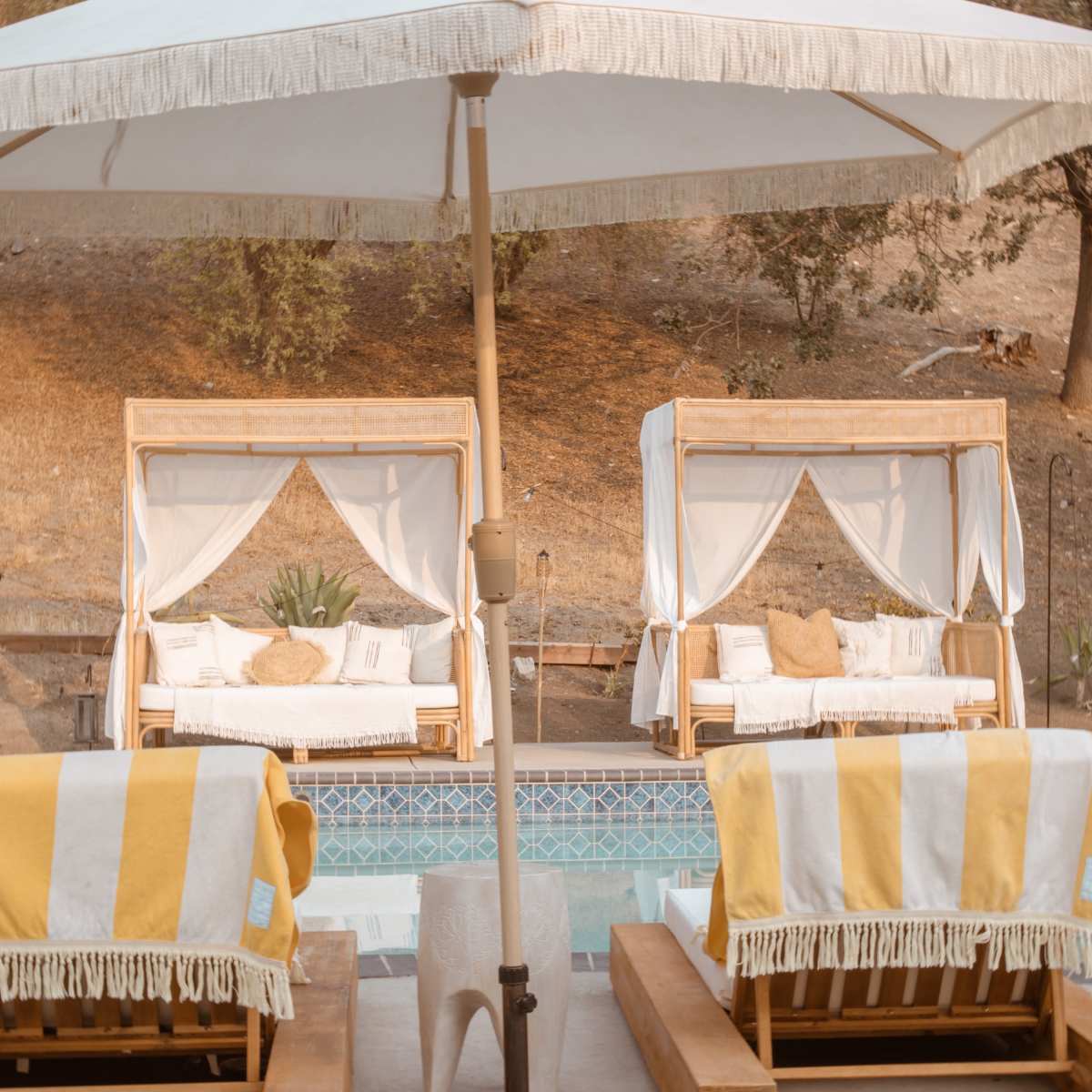 Two cabanas with white drapes beside a swimming pool, accompanied by yellow and white striped towels on lounge chairs underneath a large umbrella.