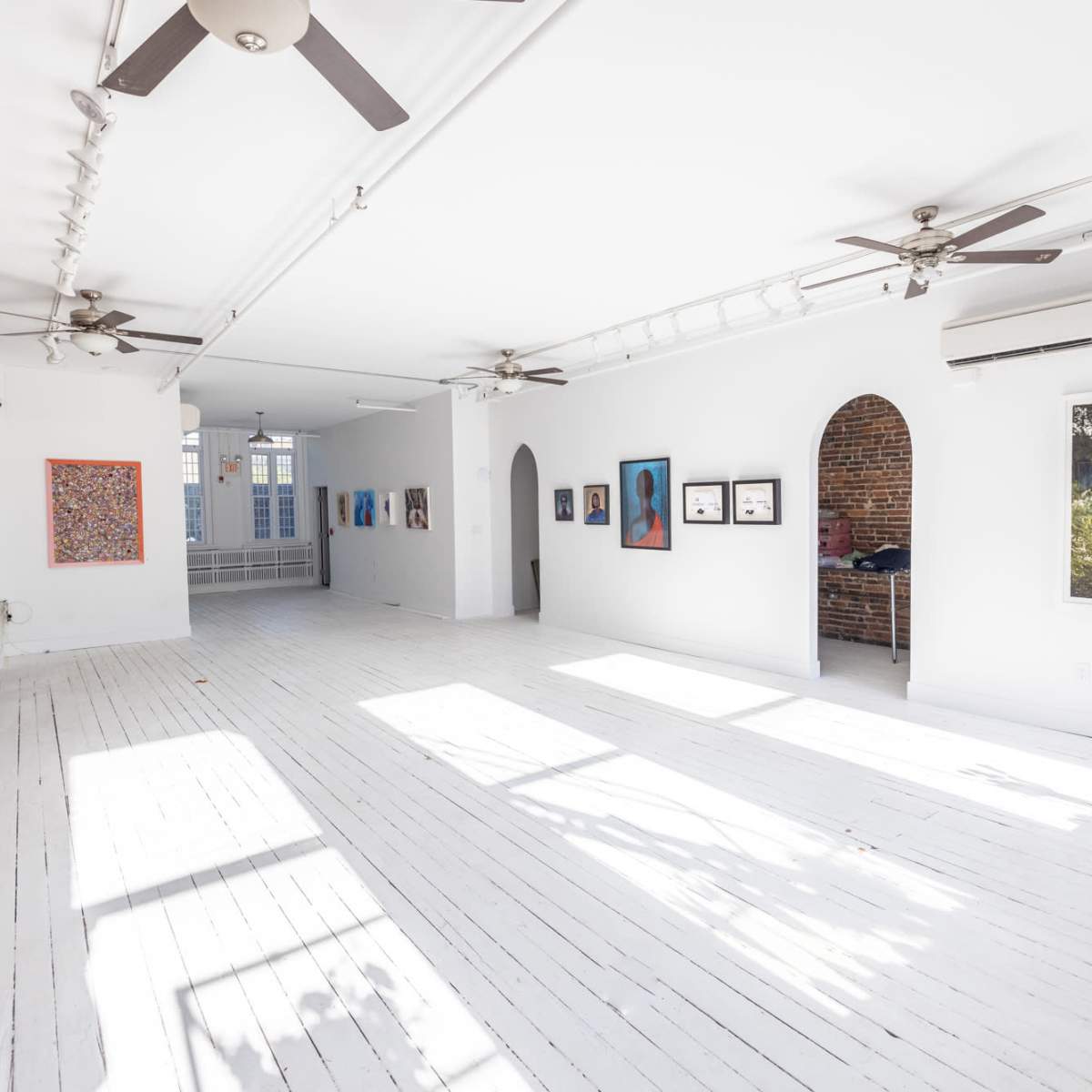A spacious art gallery with white wooden floors, exposed brick walls, and various framed artworks hung on the walls.