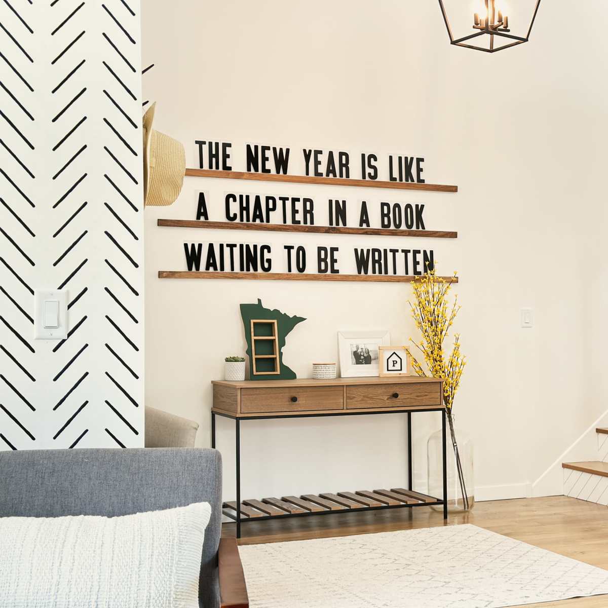 A modern living room with a decorative wall featuring wooden shelves that display the phrase "THE NEW YEAR IS LIKE A CHAPTER IN A BOOK WAITING TO BE WRITTEN" alongside a staircase and a tabletop decor setup.