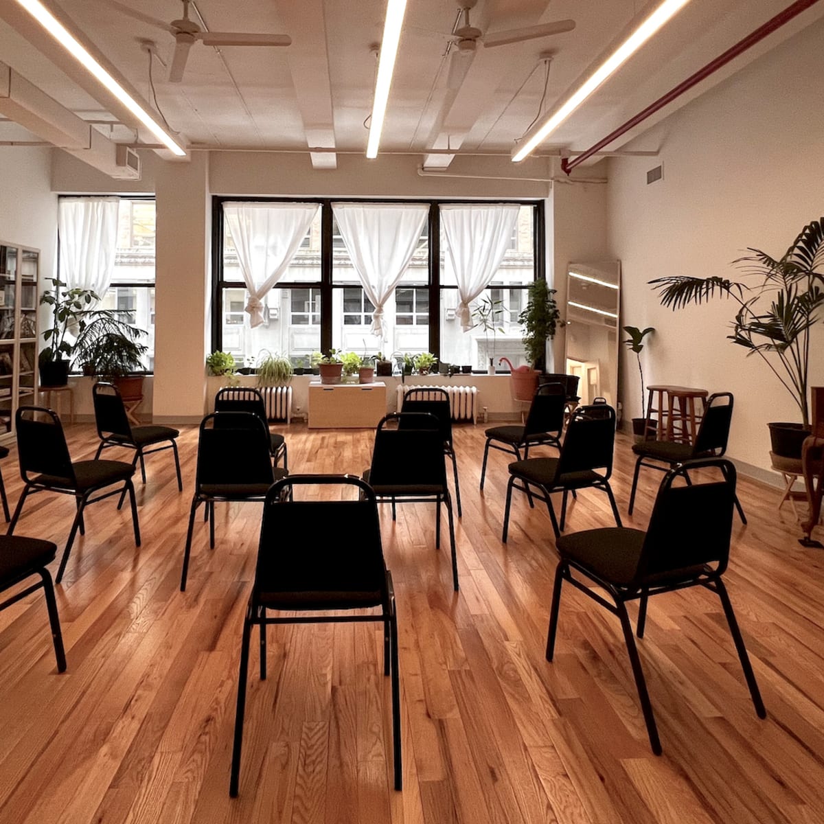 A spacious room with hardwood floors contains circular seating arranged with black chairs around a central table, illuminated by natural light from large windows.