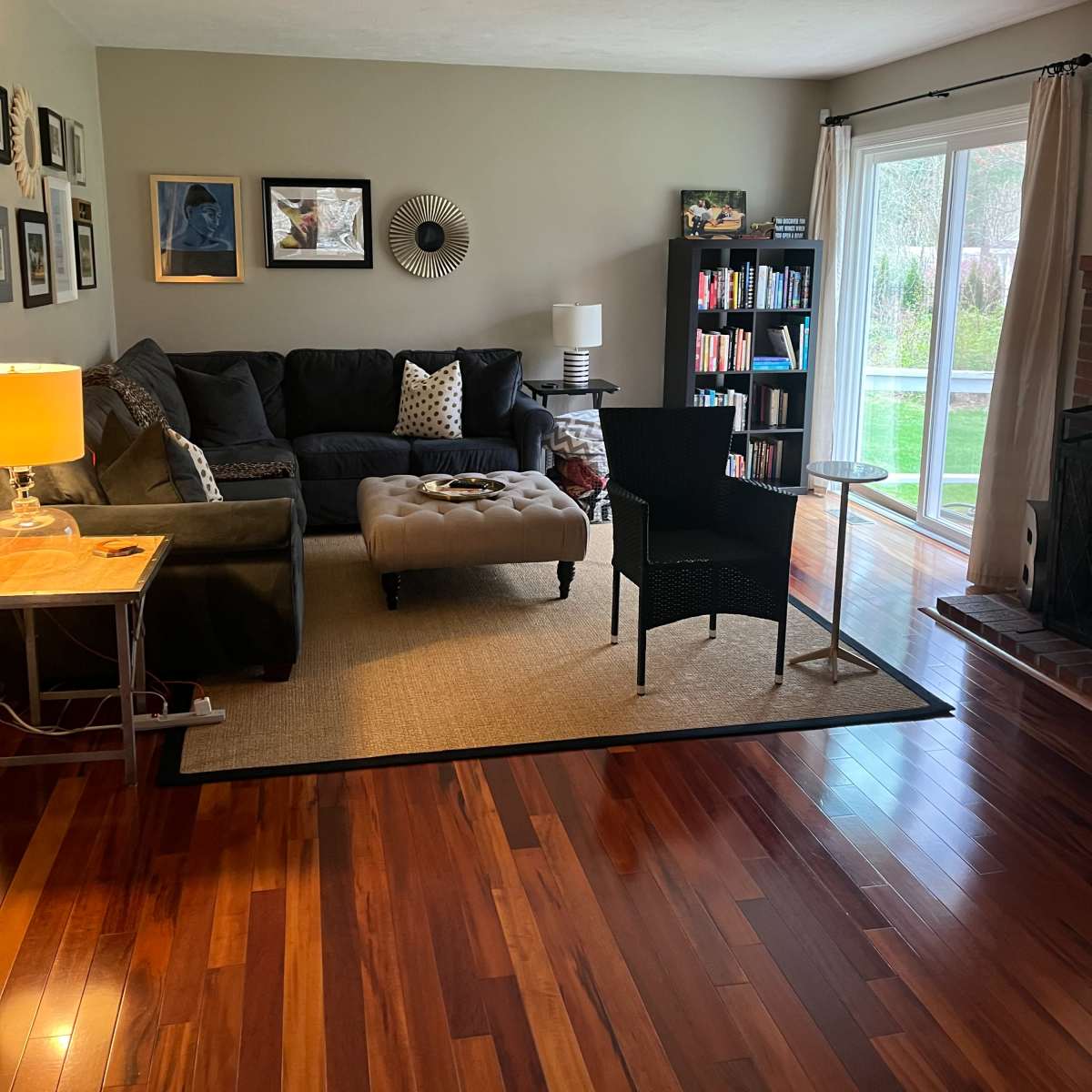The living room features a dark sofa, an ottoman, a small table with a lamp, two chairs, and a bookshelf, all set against wooden flooring and large windows overlooking a garden.
