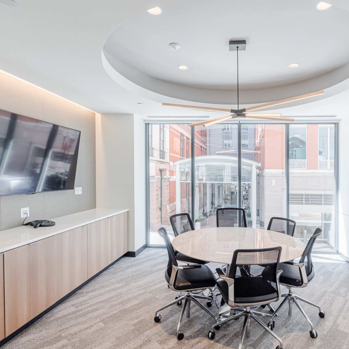 A modern conference room features a round table surrounded by six black chairs, with a large screen on the wall and windows offering a view of an outdoor space.