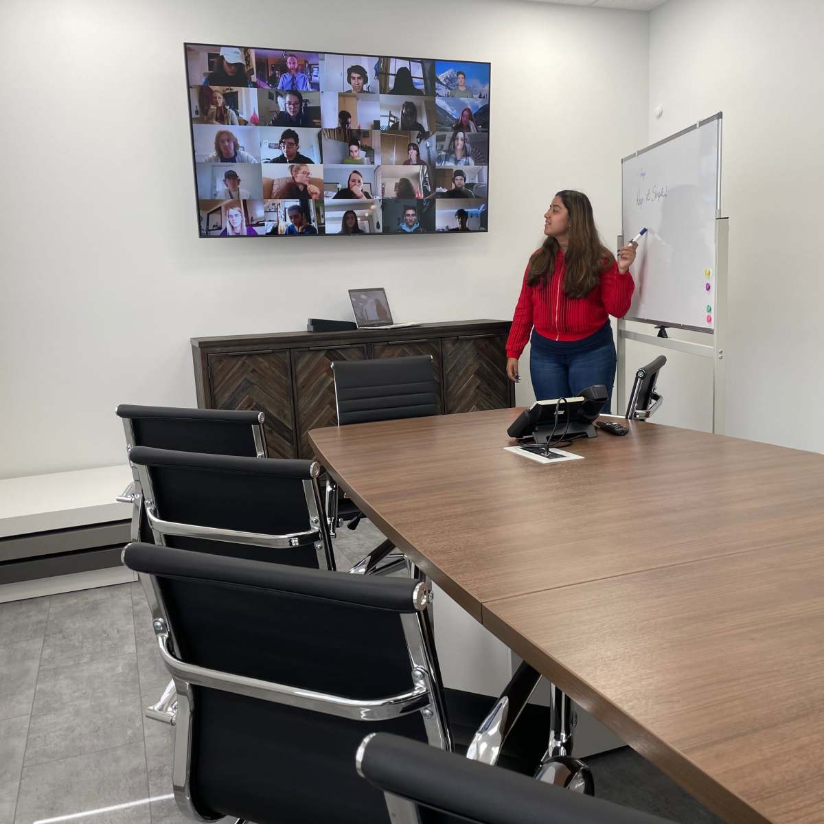 A woman presents in a conference room with a large screen displaying a virtual meeting of multiple participants.