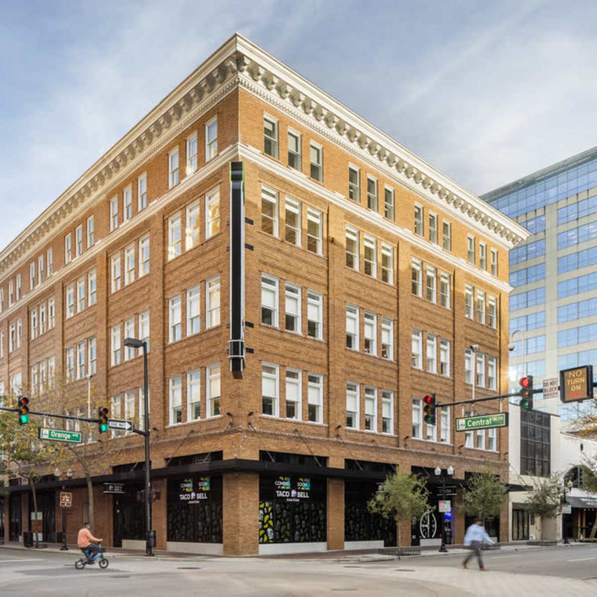 A five-story brick building with numerous windows stands at the intersection of two streets, with a modern glass building visible in the background.