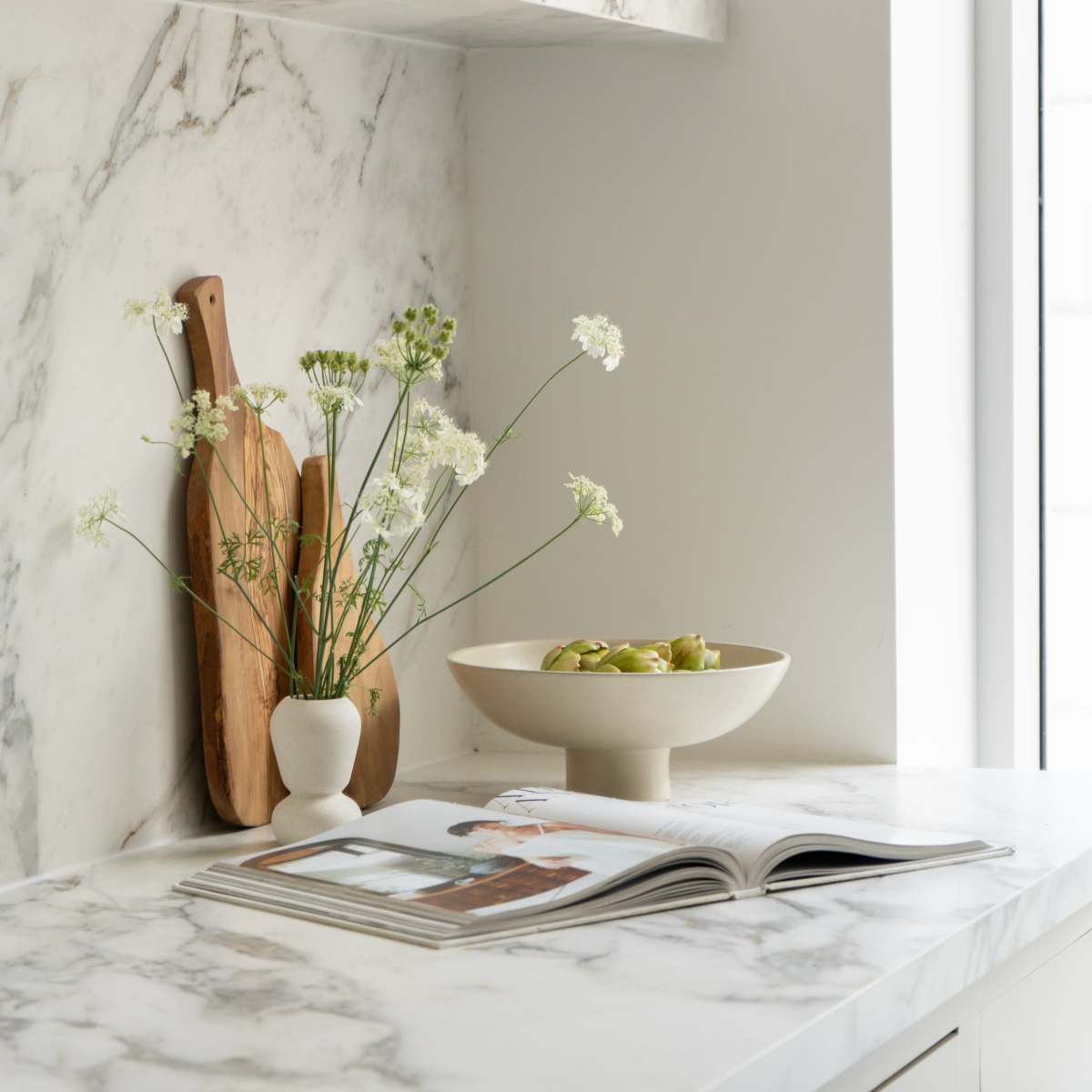 A marble countertop features a bowl of green olives, a vase of white flowers, cutting boards, and a magazine.