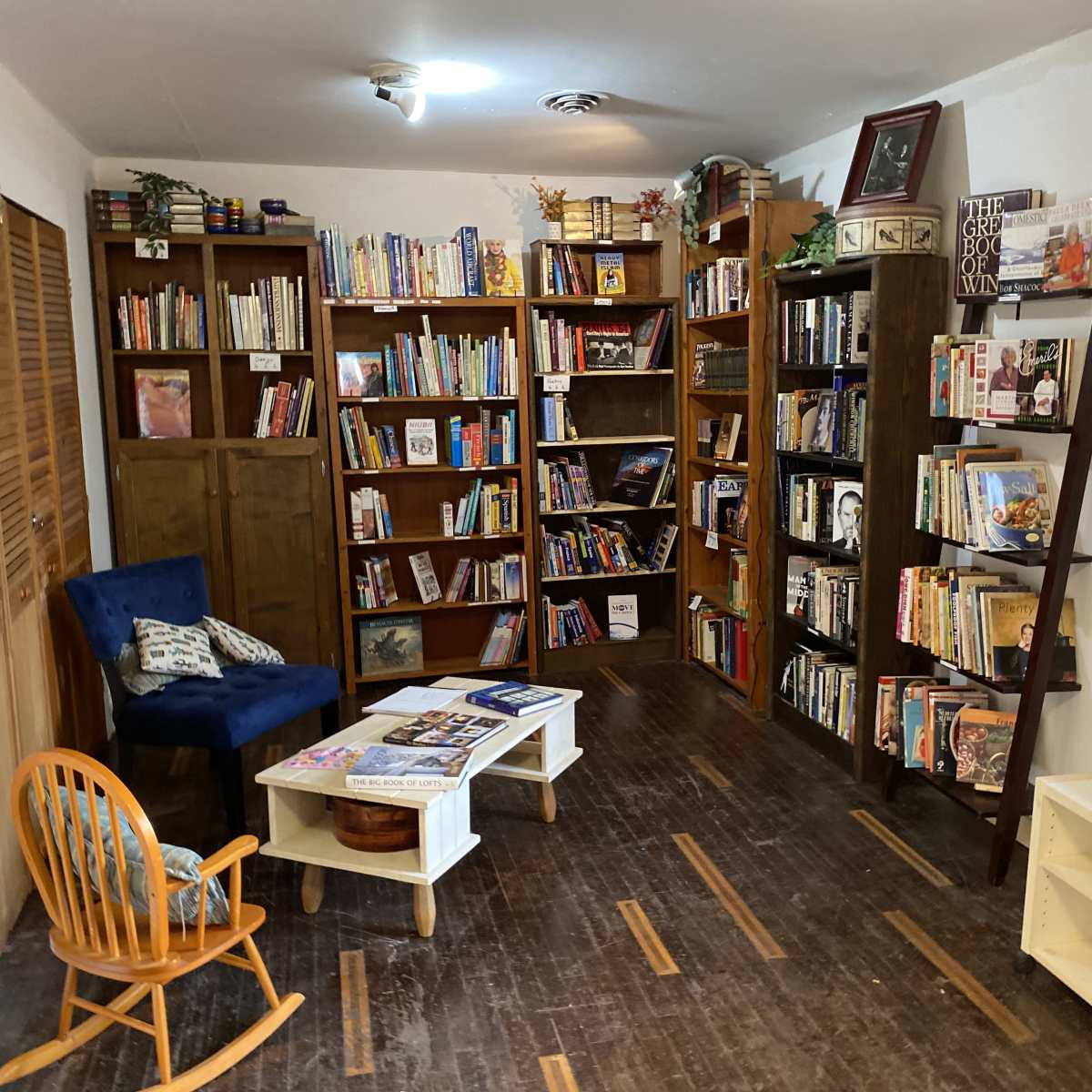 A cozy reading nook with bookshelves filled with various books, a small table, a blue chair, and a wooden ladder shelf in a well-lit room.