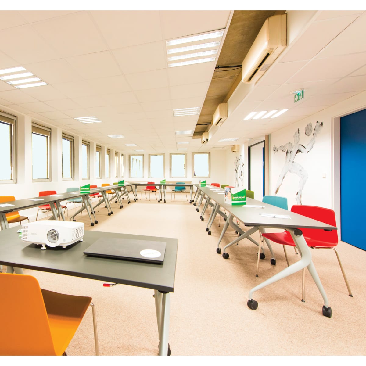 A bright, modern classroom features multiple tables arranged in a U-shape with colorful chairs and a projector in the center.