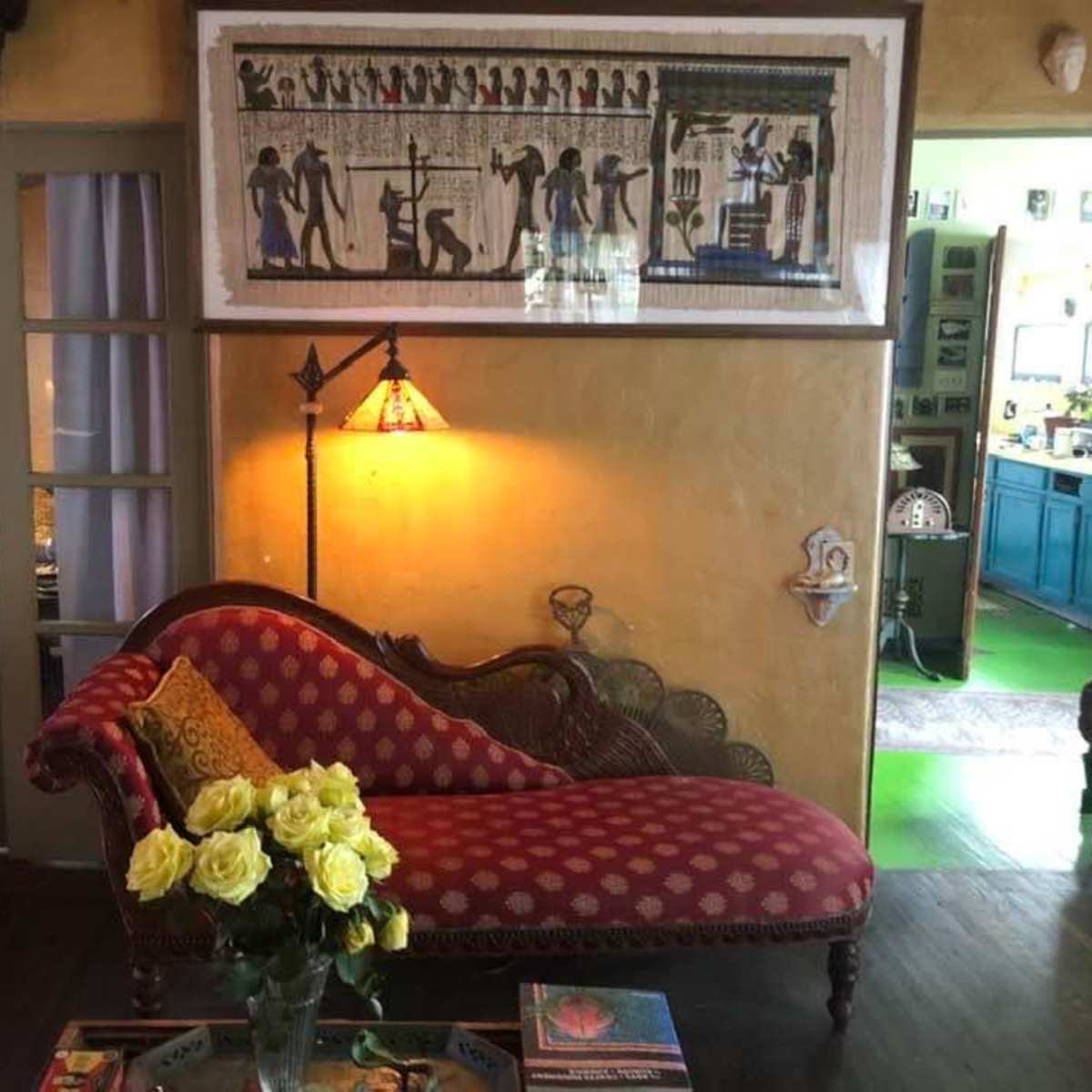 A living room with a red patterned chaise lounge, a floral arrangement of yellow roses on a coffee table, and a wall adorned with an Egyptian-themed artwork.