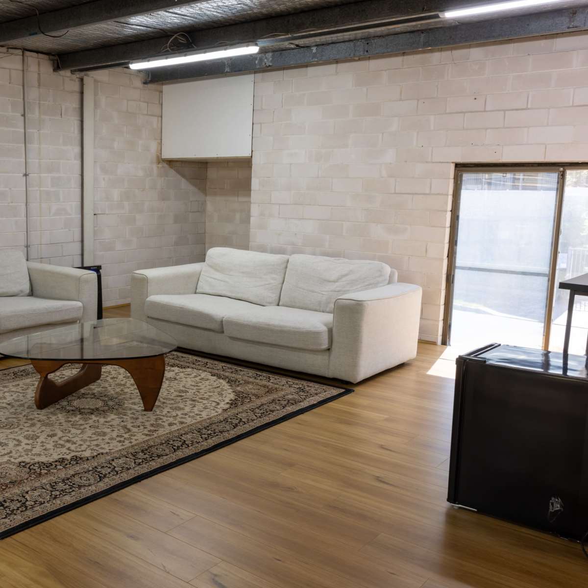 A spacious room features two light-colored sofas facing a glass coffee table on a patterned rug, with a large window allowing natural light.