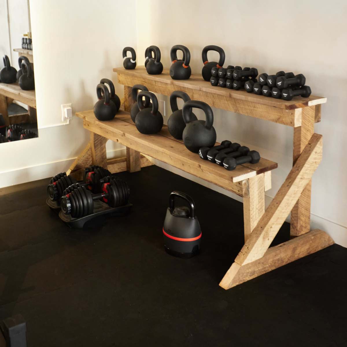 A wooden rack displays an assortment of black kettlebells and dumbbells in a gym setting.