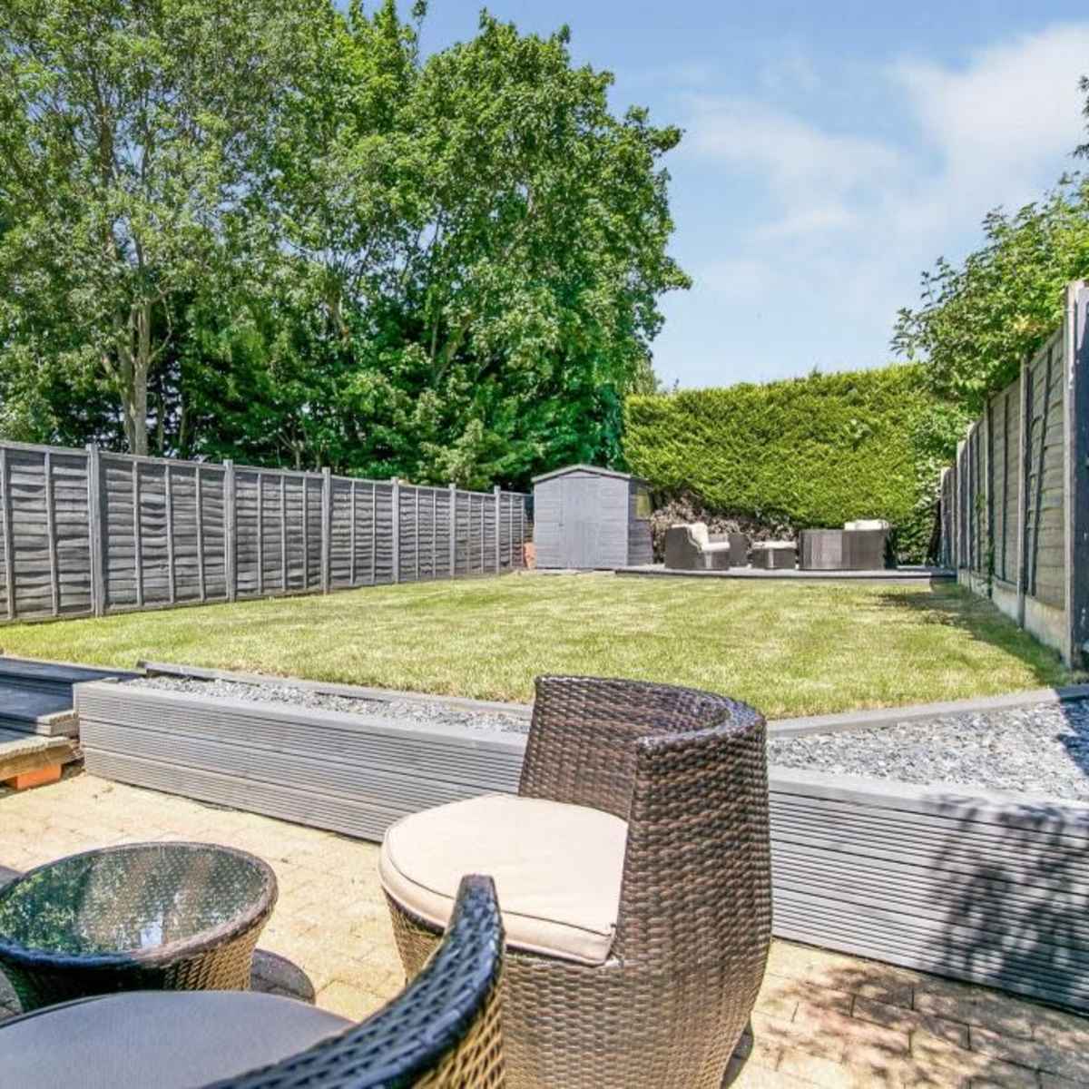 A landscaped garden with a grassy area, a wooden deck, and a stone pathway, surrounded by wooden fences and trees.