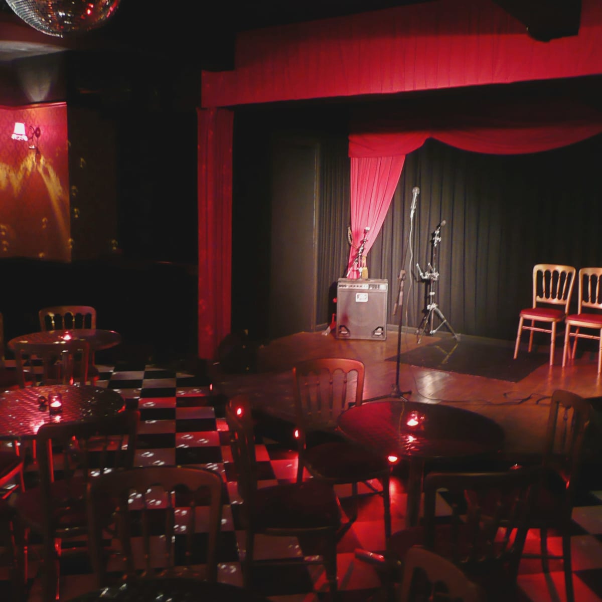 A small music venue with a stage featuring red curtains, two microphones, and three empty chairs, surrounded by tables and chairs on a checkered floor.