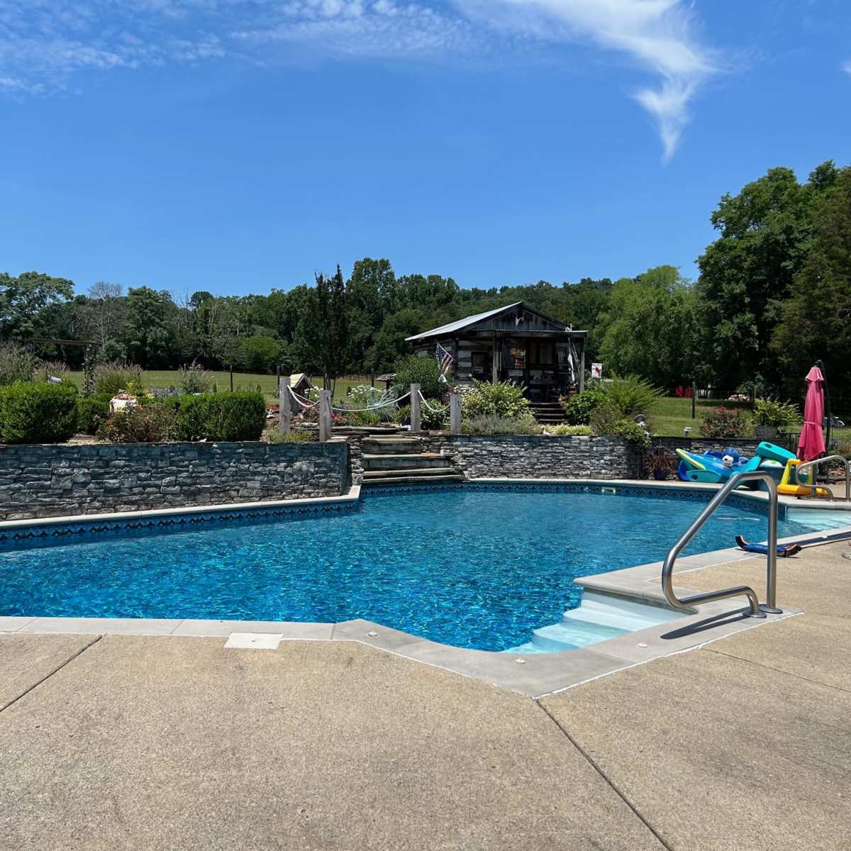 A clear blue swimming pool surrounded by a concrete deck, with a stone wall and a grassy area in the background.