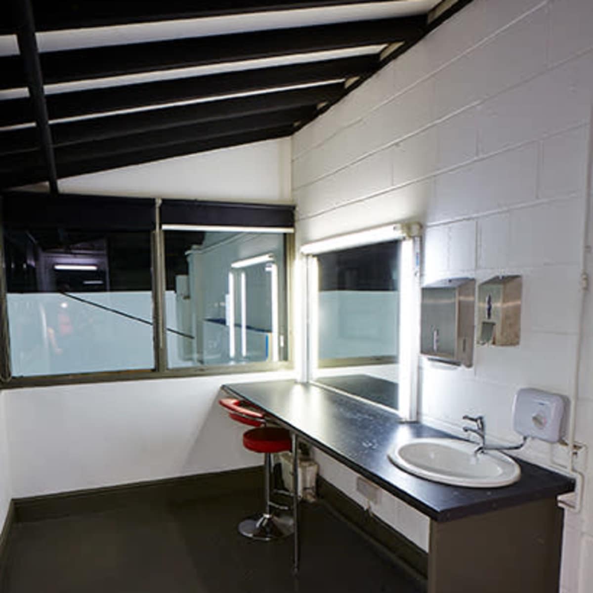 A minimalist restroom with a sink, a red stool, and a large mirror illuminated by fluorescent lights.