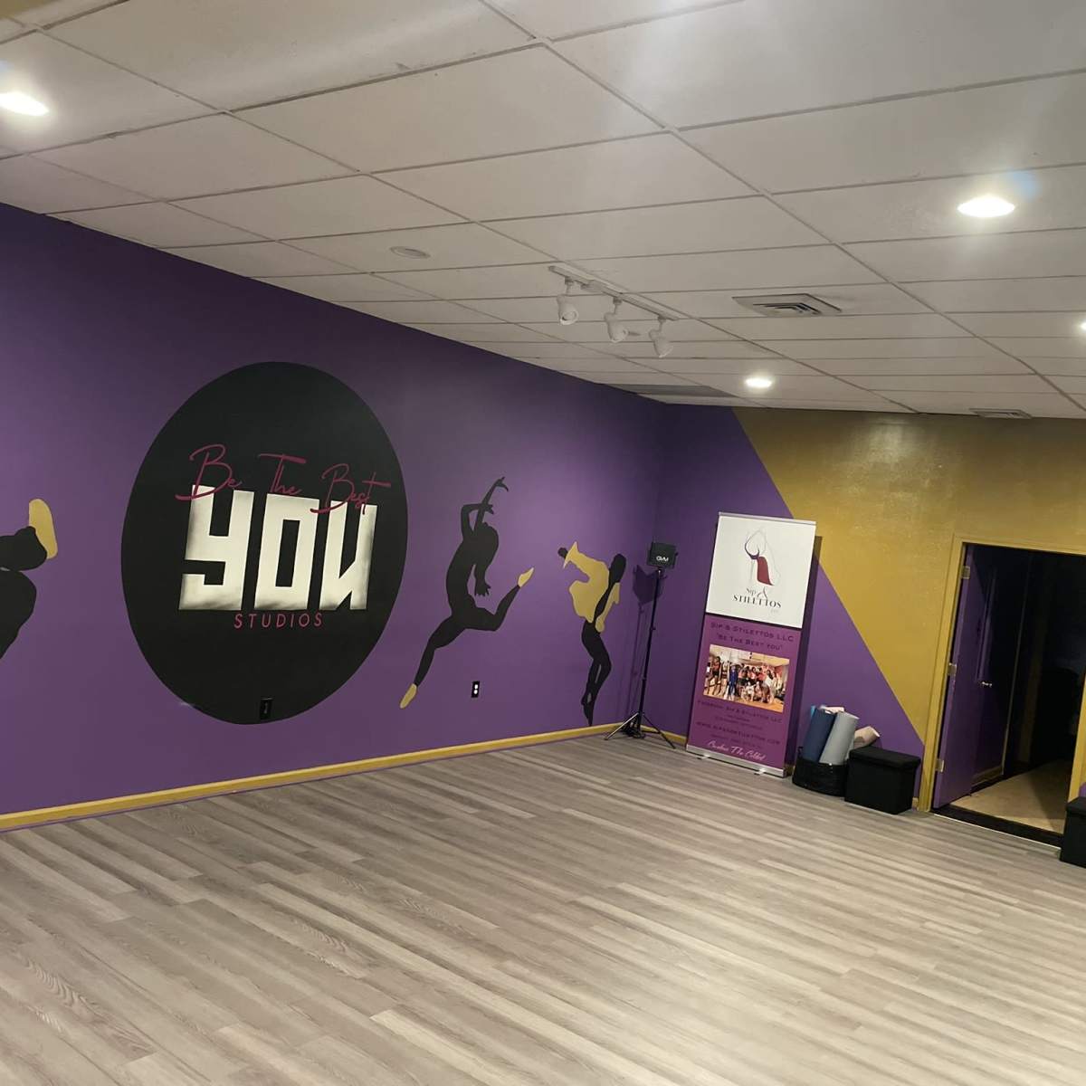 A dance studio with a purple wall featuring a circular logo that reads "Be The Bit YOU Studios," and a yellow accent wall with an entrance to another room.