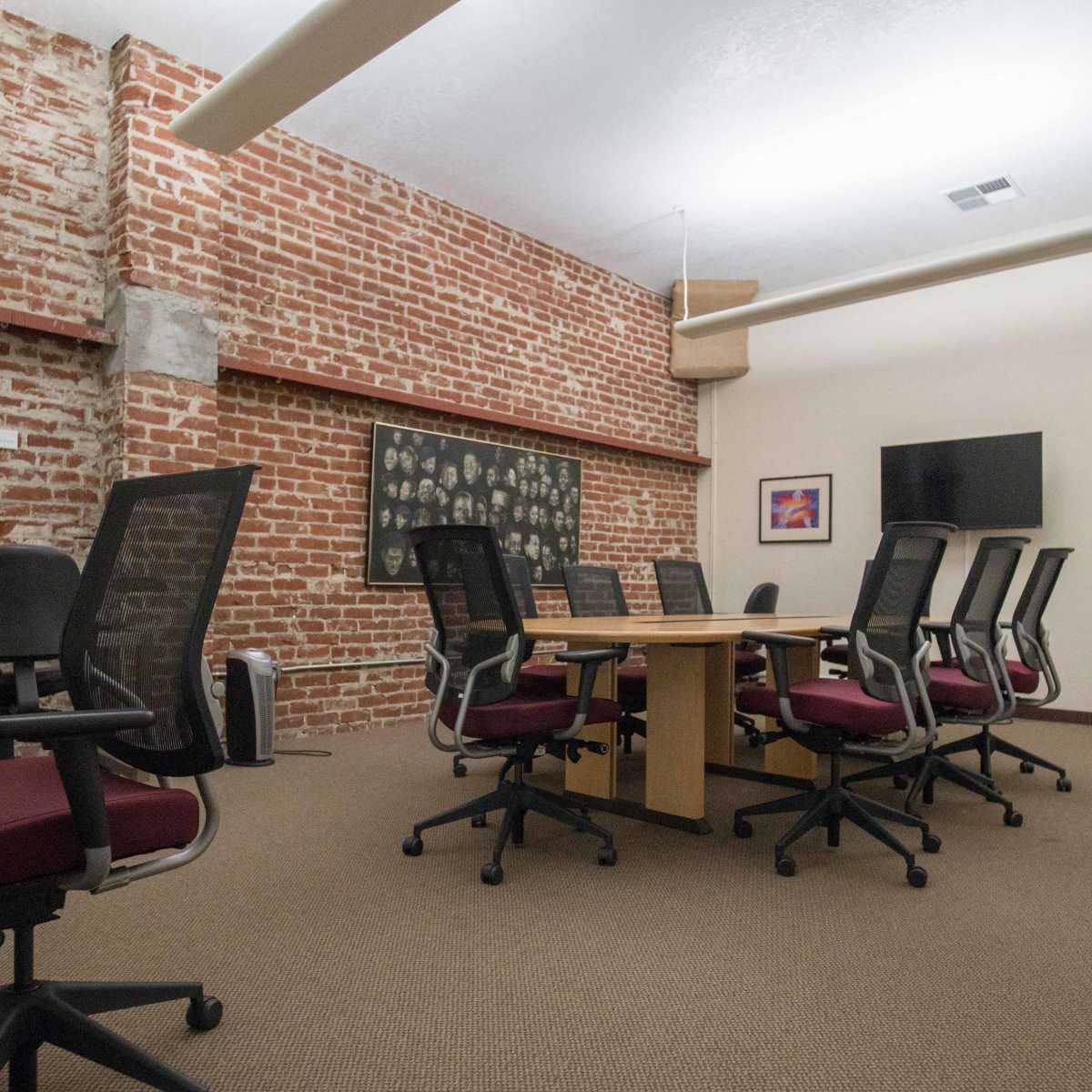 A conference room features a wooden table surrounded by office chairs, with brick walls and framed artwork.