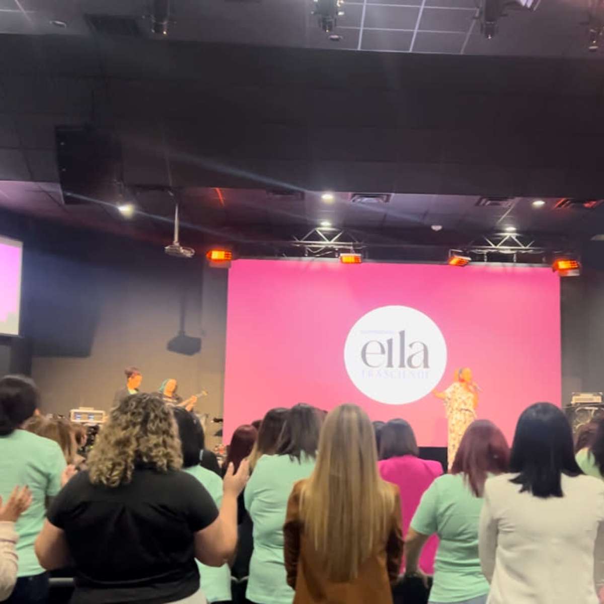 A crowd of women stands with hands raised in a dimly lit venue, facing a stage where a speaker addresses them, with a large screen displaying the word "ella."