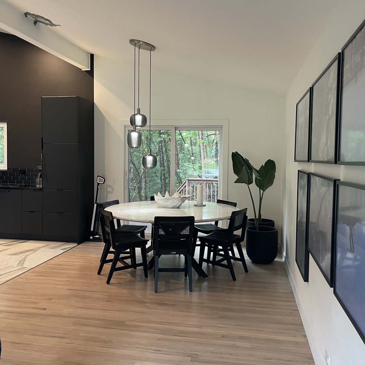 A modern dining area features a round table surrounded by black chairs, with large windows overlooking a wooded view and framed artworks on the wall.