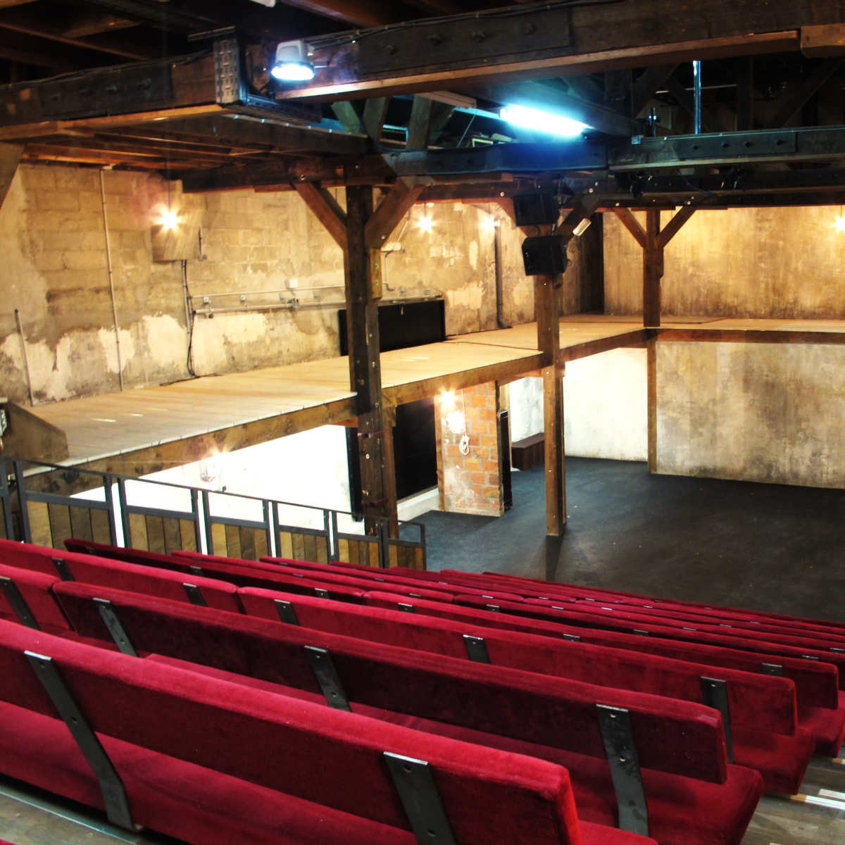 A spacious, empty theater with wooden beams and red seating arranged in tiers facing a simple stage area.